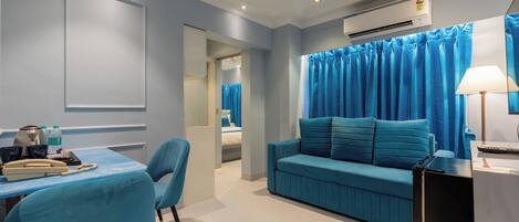 A modern hotel room with a blue sofa, a desk with a telephone and a lamp, and an air conditioning unit on the wall.