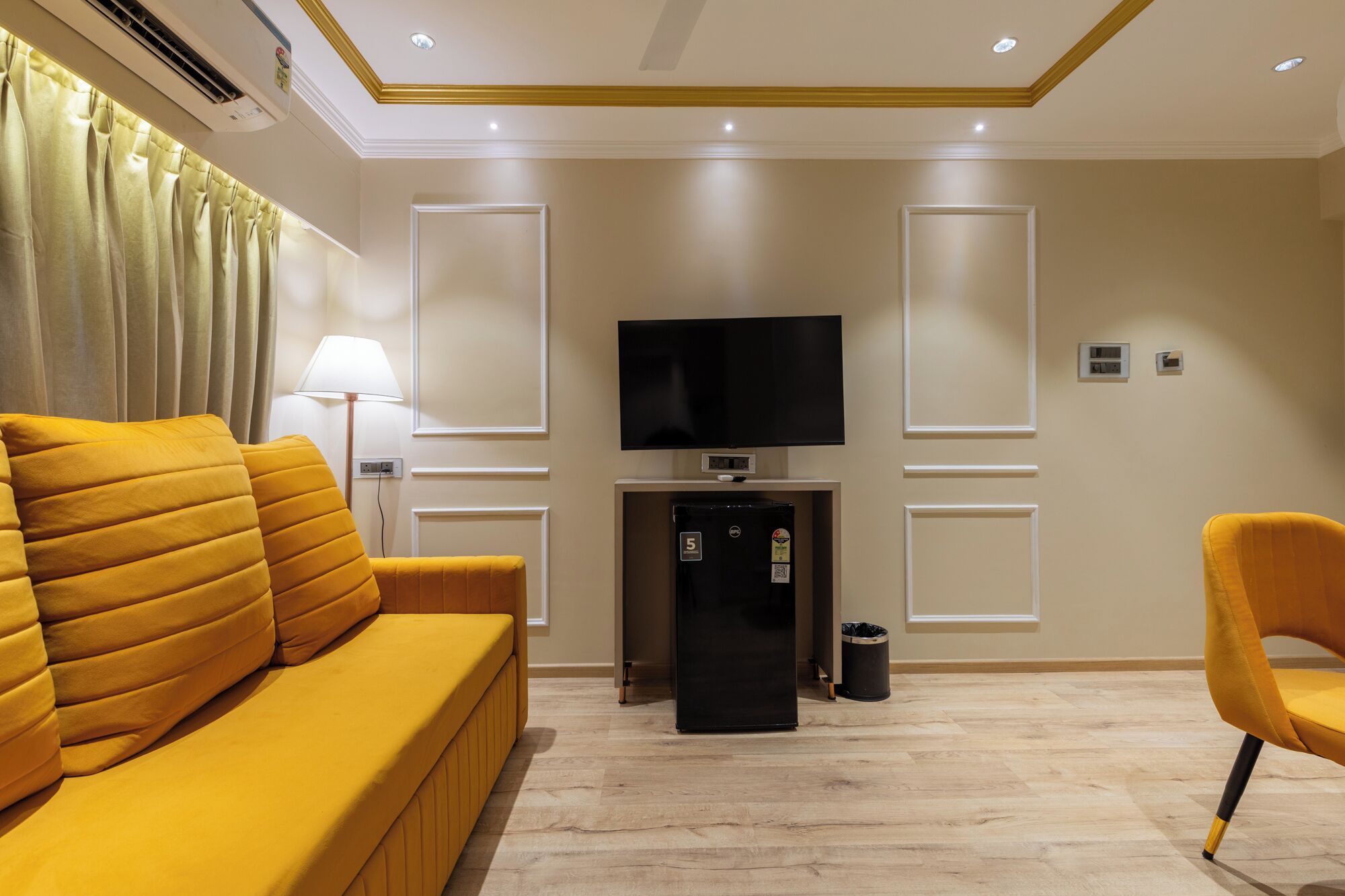 A modern living room with a yellow sofa, a black refrigerator, a flat-screen TV, and a lamp.