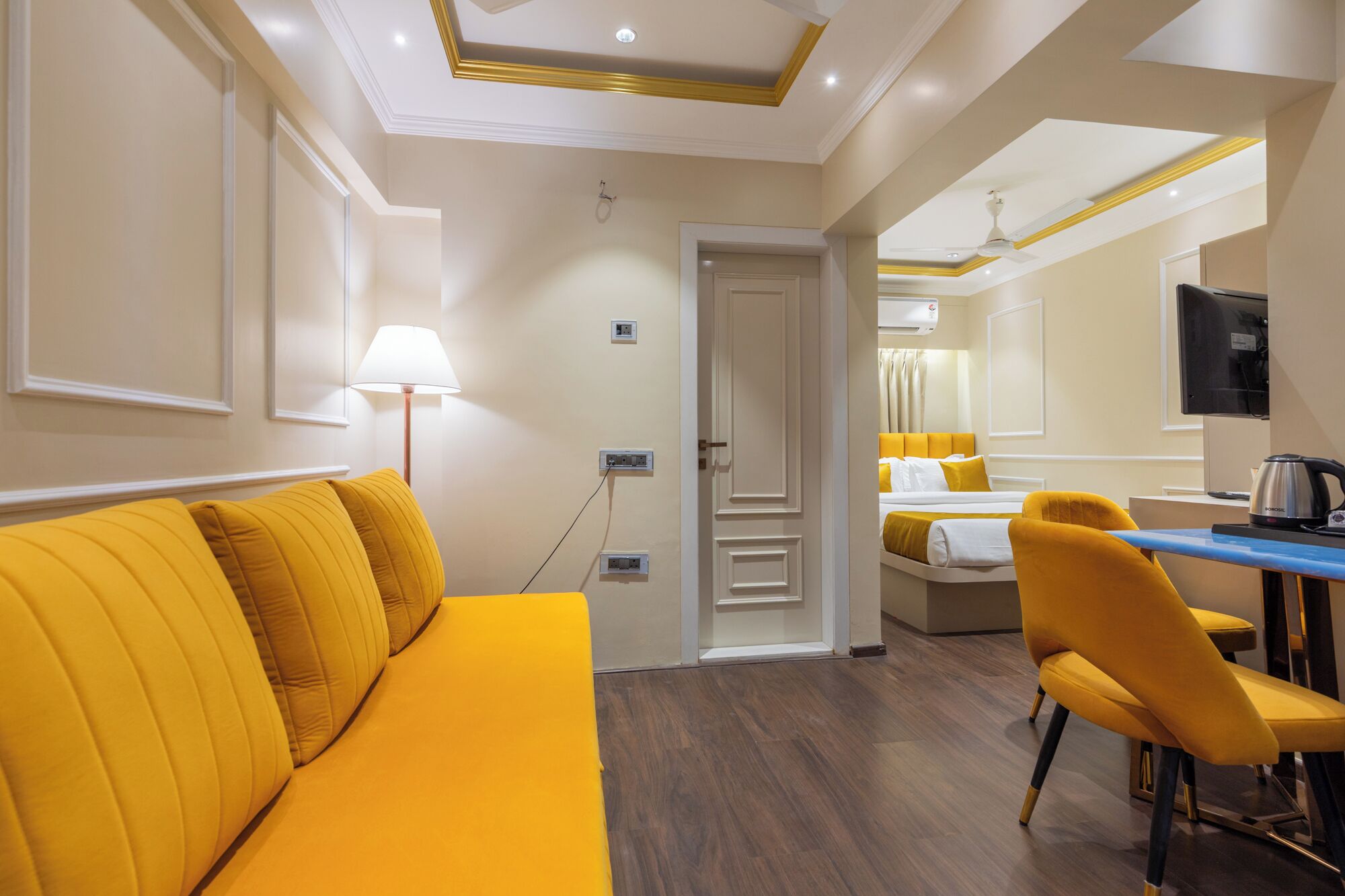 Habitación de hotel con sofá amarillo, un escritorio con silla, cama con ropa de cama amarilla y televisión en la pared.