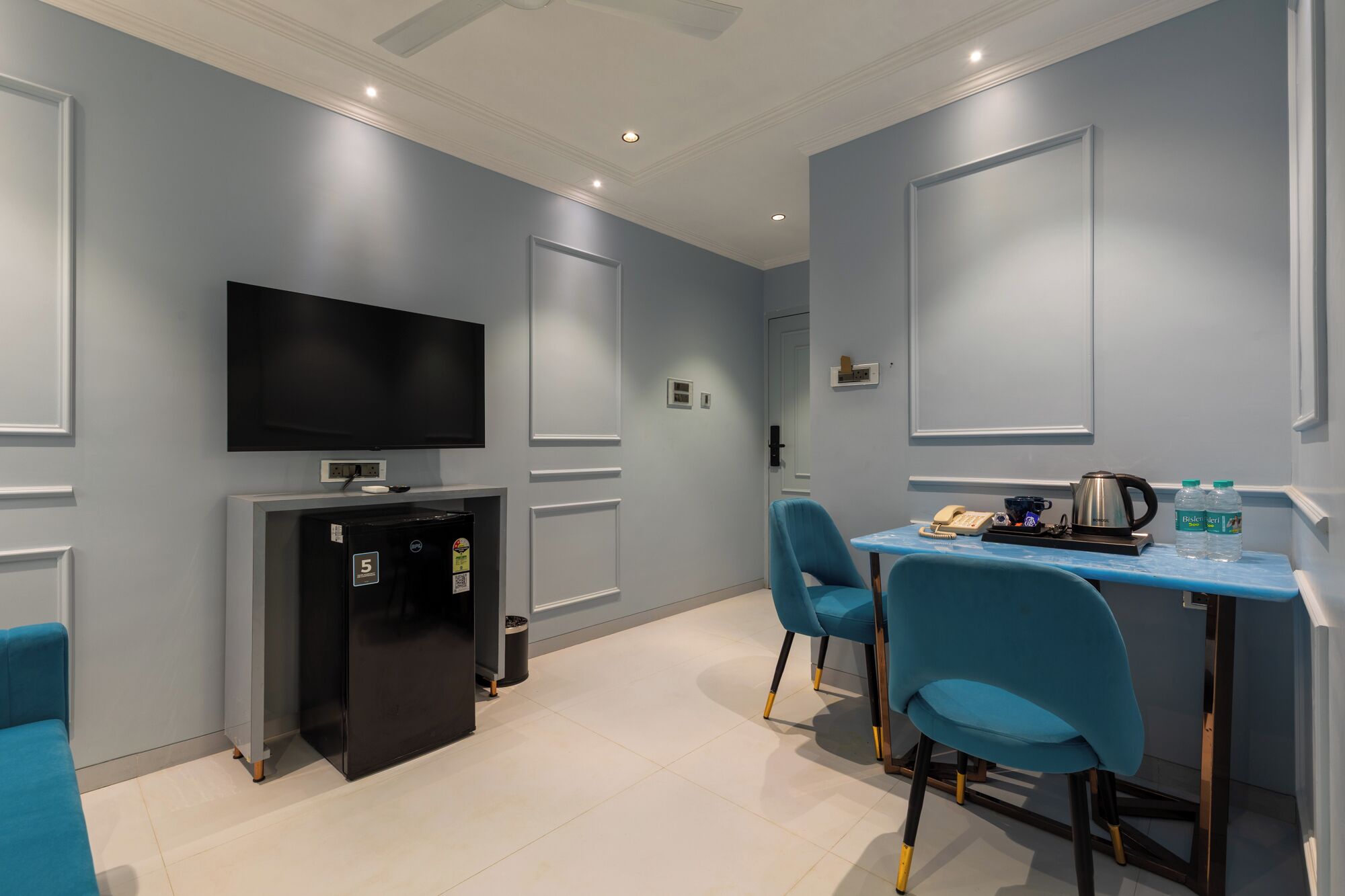 A modern hotel room with a flat-screen TV, a minibar, a coffee machine, and two blue chairs.