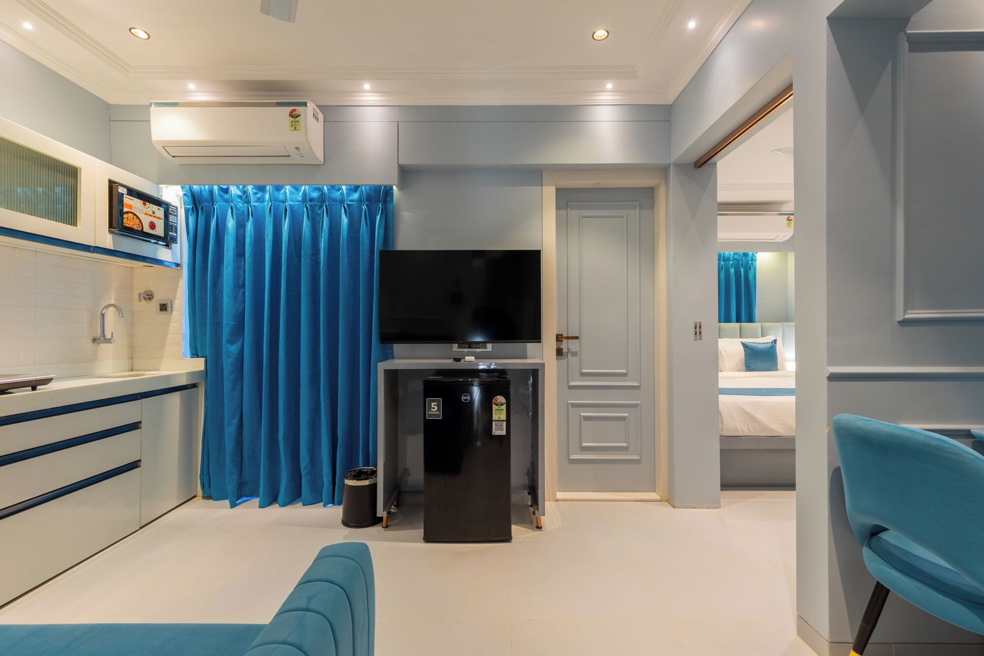 A compact hotel room with a kitchenette, a flat-screen TV, a refrigerator, and a blue chair.