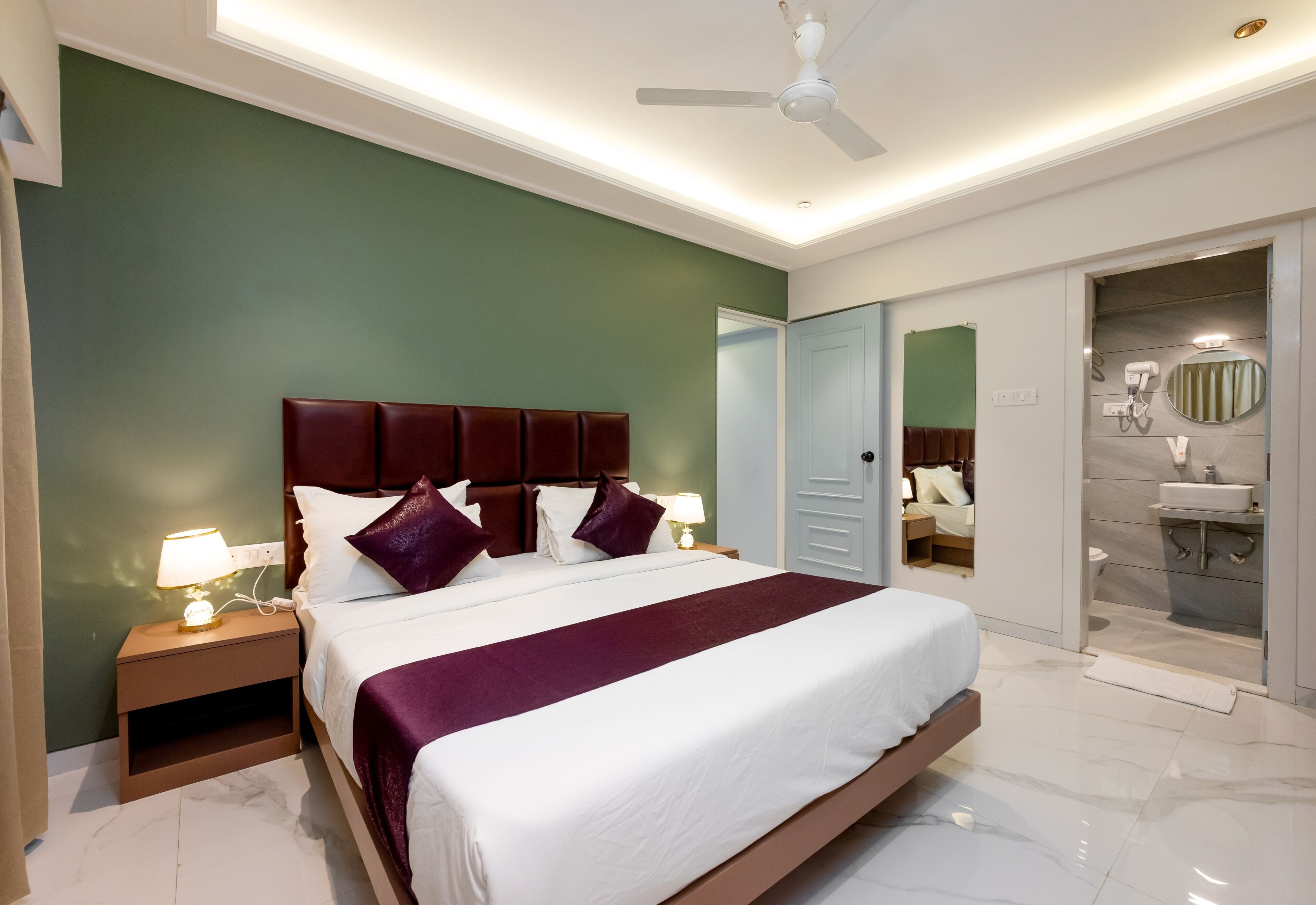 A hotel room with a large bed, a nightstand, a lamp, a ceiling fan, and a bathroom visible through an open door.