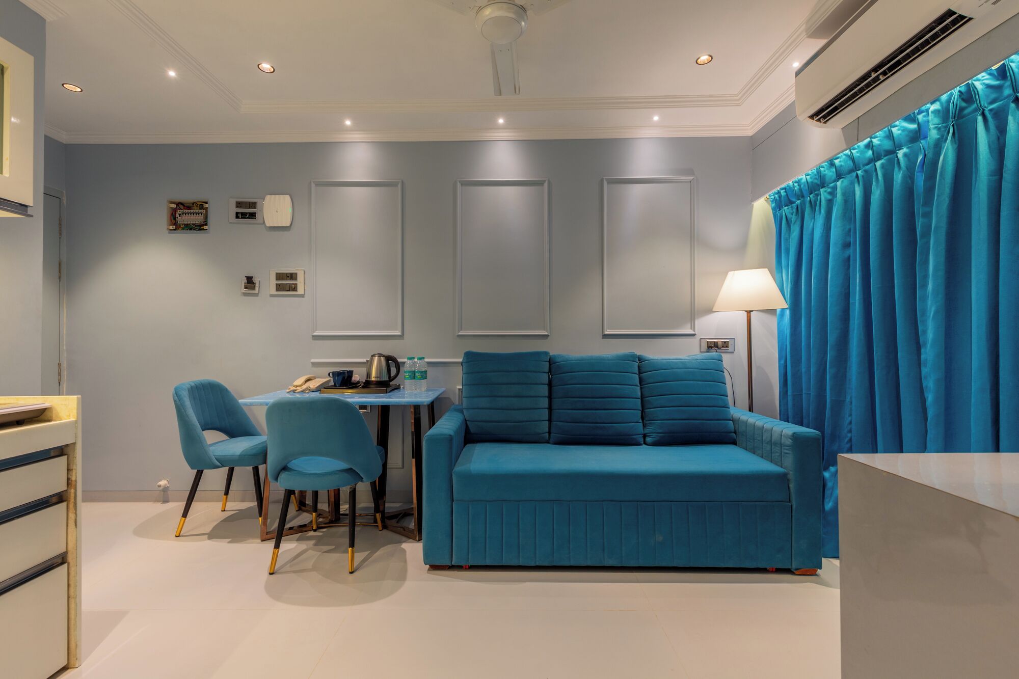 A room with a blue sofa, two matching chairs, a small table with a kettle and cups, and a lamp.