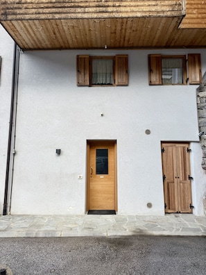 A two-story building with a wooden door and windows, a stone wall, and a metal gutter.