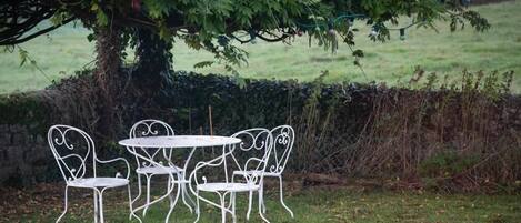 A white metal table and chairs set under a tree with a hedge in the background.
