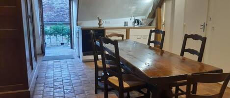 A dining area with a wooden table and chairs, a kitchen in the background, and a view of the outside through an open door.