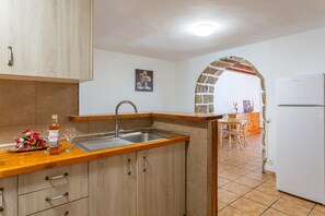 Eine Küche mit Holzschränken, einem Spülbecken, einer Arbeitsplatte mit Flasche und Glas sowie einem steinernen Bogen, der in einen Essbereich führt.