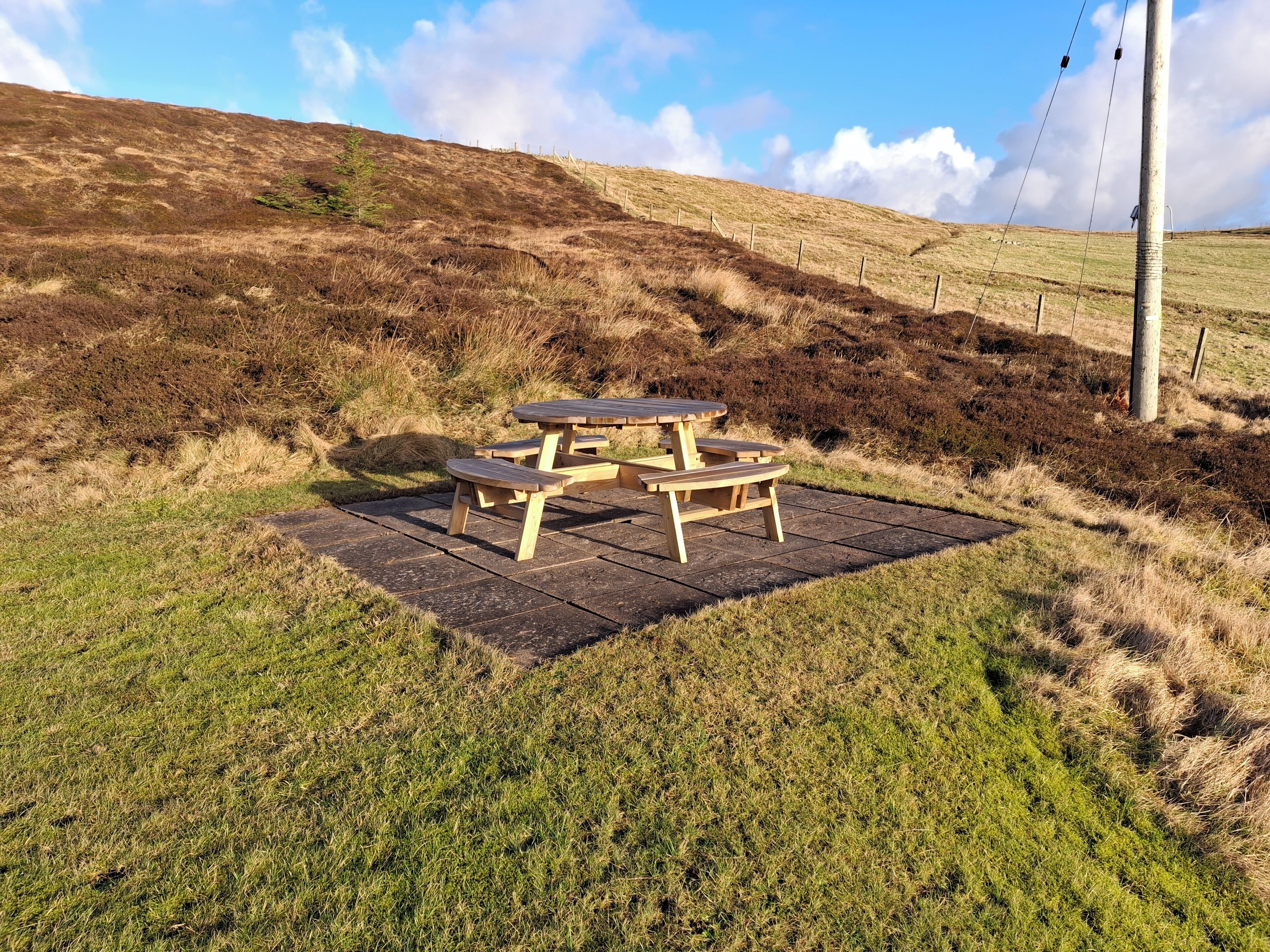 Et picnicbord placeret på en græsplæne med et bakket landskab i baggrunden.