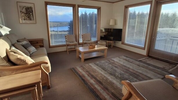 Een ruime woonkamer met een bank, stoelen, een salontafel en grote ramen met uitzicht op het omliggende landschap.