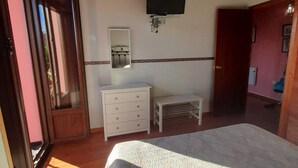 Ein Schlafzimmer mit einem Bett, einem Schminktisch, einem Spiegel, einem an der Wand montierten Fernseher und einer Tür zu einem anderen Raum.