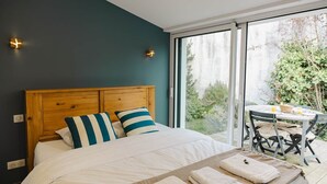 A bedroom with a wooden headboard, a bed with white bedding, two pillows, a table with chairs, and a view of greenery outside. - Kallisté - Stunning Oceanview Villa (Île-d'Aix)