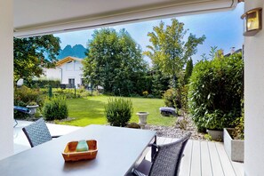 A patio with a table and chairs, a view of a garden with various plants and a building in the background. - Vacation apt. Verena in Garmisch – stylish, central, and with green garden oasis (Garmisch-Partenkirchen)