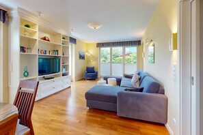 A modern living room with a flat-screen TV, a grey sofa, a dining table with chairs, and a bookshelf. - Vacation apt. Verena in Garmisch – stylish, central, and with green garden oasis (Garmisch-Partenkirchen)