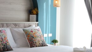 A neatly made bed with floral pillows, a bedside plant, and a hanging light fixture in a room with a blue accent wall and a large window with curtains. - J The Park Condotel (Hat Yai)