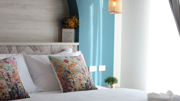 A neatly made bed with floral pillows, a small plant, and a hanging light fixture in a room with teal walls and a white headboard. - J The Park Condotel (Hat Yai)