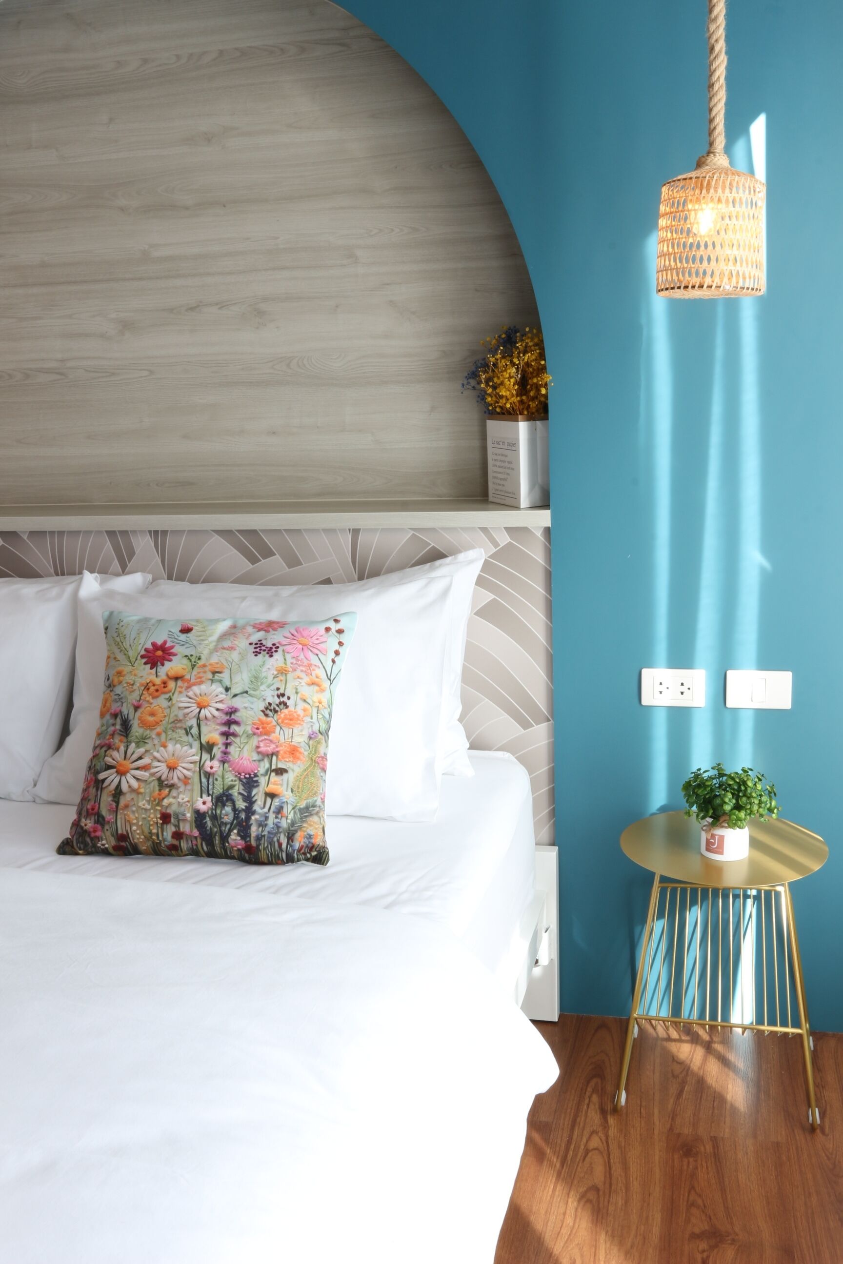 Een slaapkamer met een gebogen hoofdeinde, een bloemenkussen, een nachtkastje met een plant en een hangende lamp.