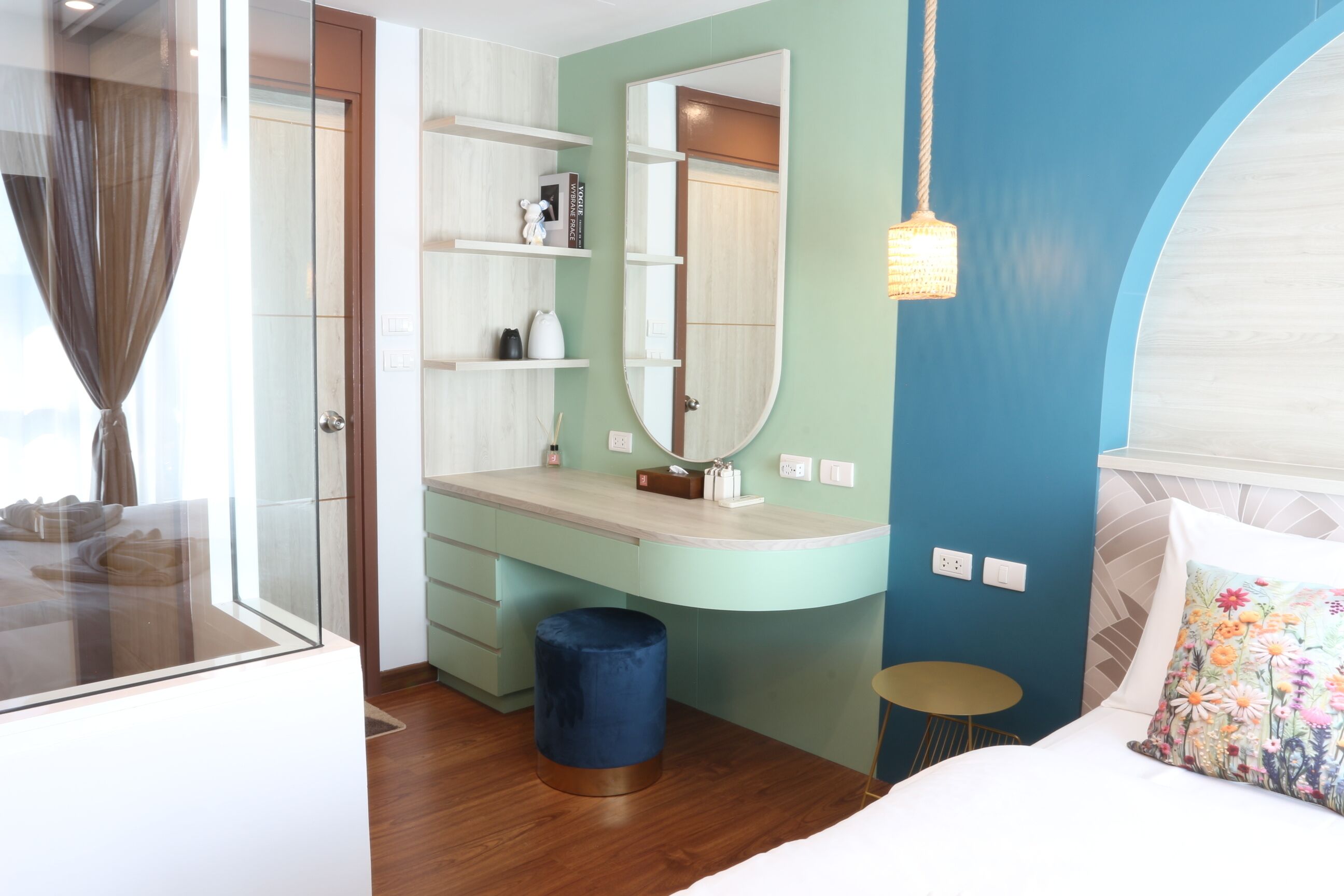 A modern bedroom with a large bed, a vanity with a mirror, a small stool, and a blue trash bin.