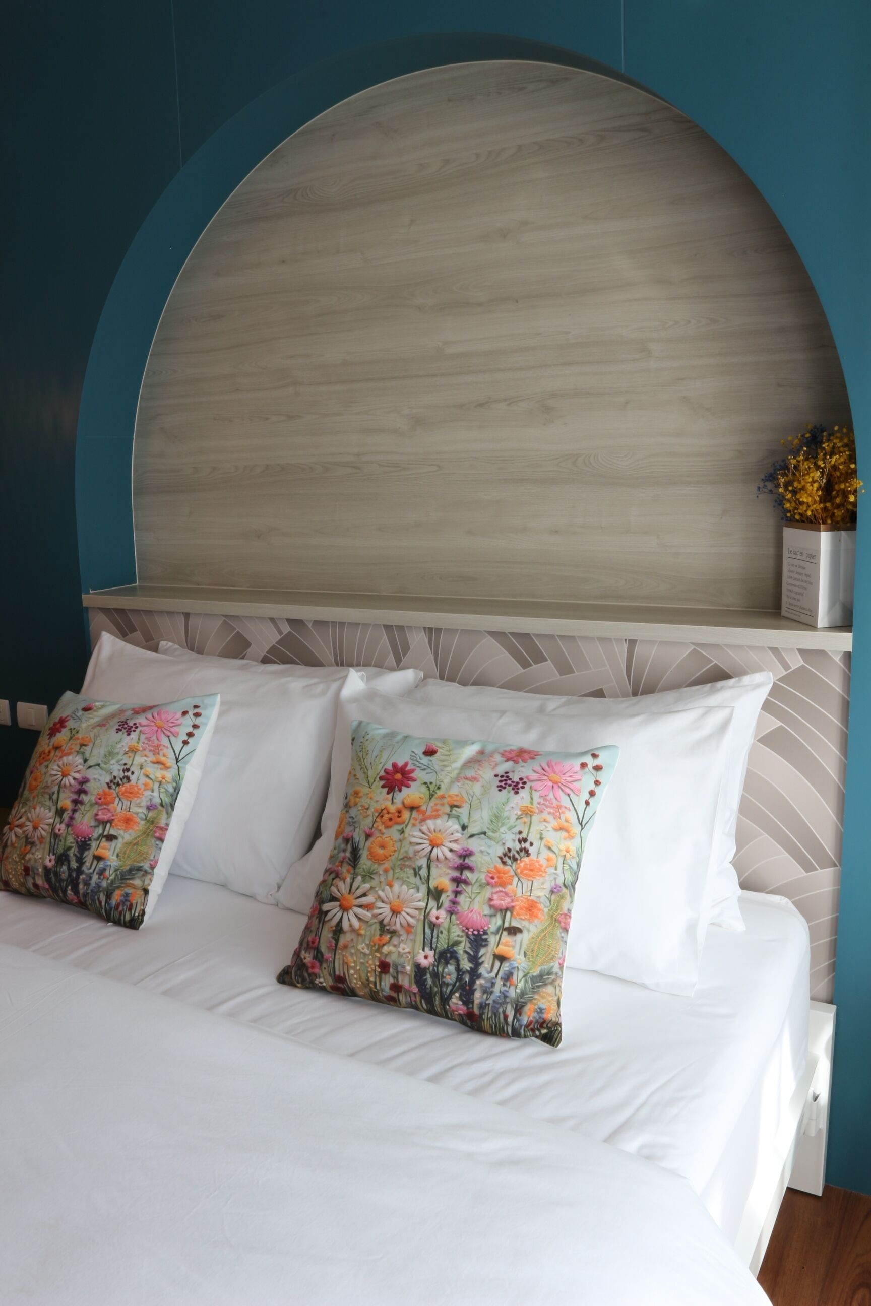 Een bed met witte lakens en sierkussens met bloemenprint, een houten hoofdbord en een muur in teal kleur.