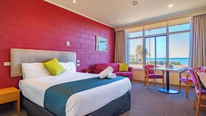 A hotel room with a large bed, a red brick wall, a window with a view, a small table with chairs, and a pink sofa. - The Beach Hotel Whyalla (Whyalla)