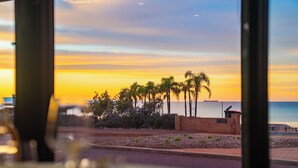 A sunset view from a window overlooking a beach with palm trees and a body of water. - The Beach Hotel Whyalla (Whyalla)