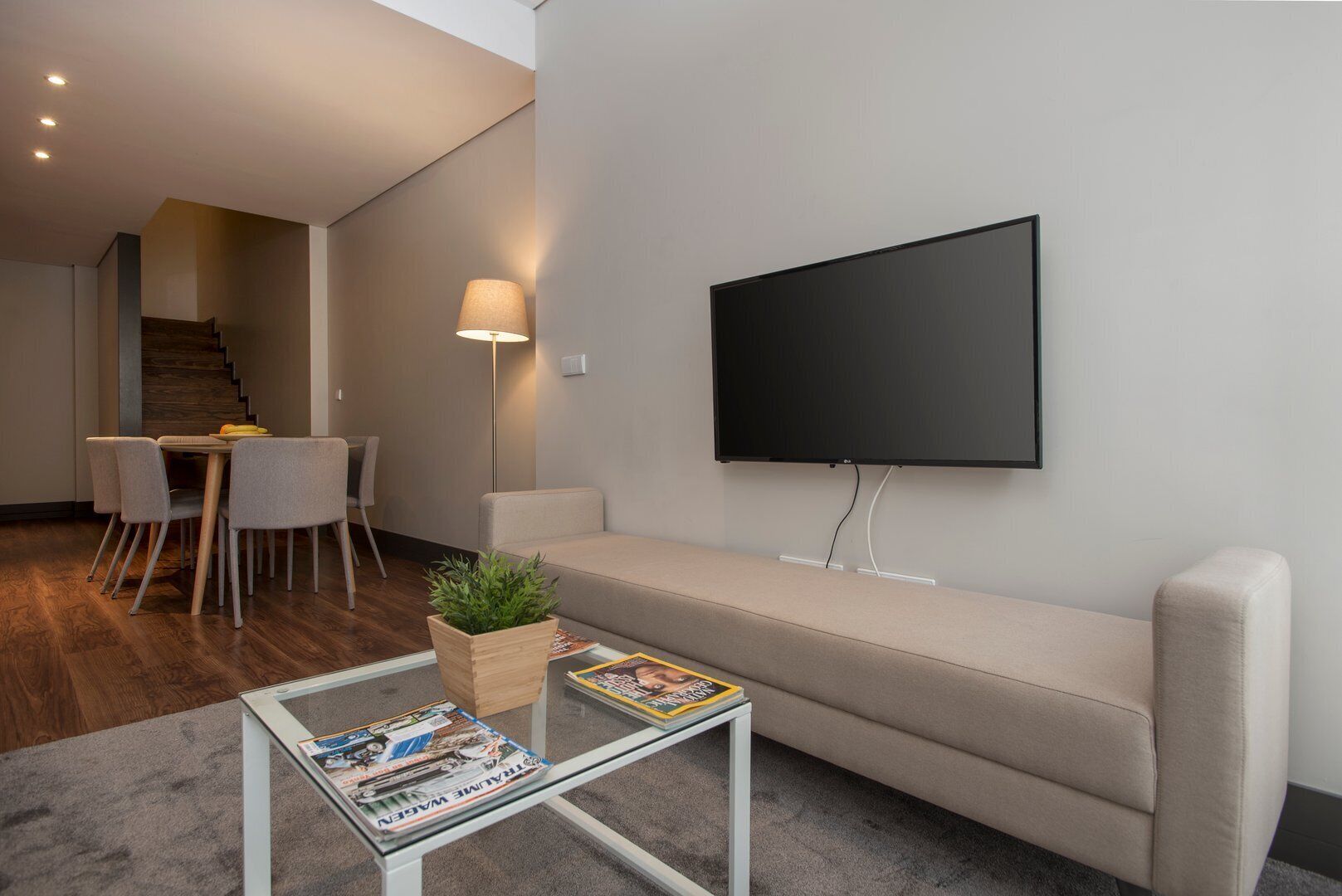 Ein modernes Wohnzimmer mit einer Couch, einem Couchtisch mit Zeitschriften, einem an der Wand montierten Fernseher und einem Essbereich mit Stühlen und einem Tisch.