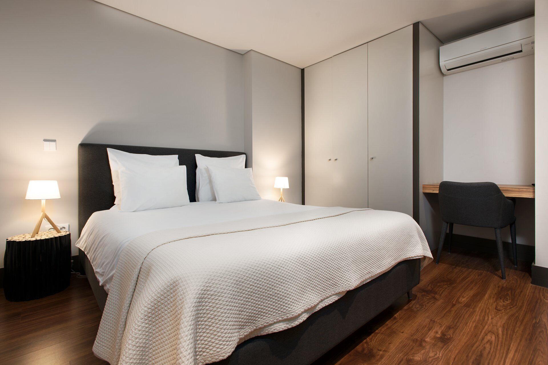 A hotel room with a bed, bedside lamps, a desk, and an air conditioning unit.