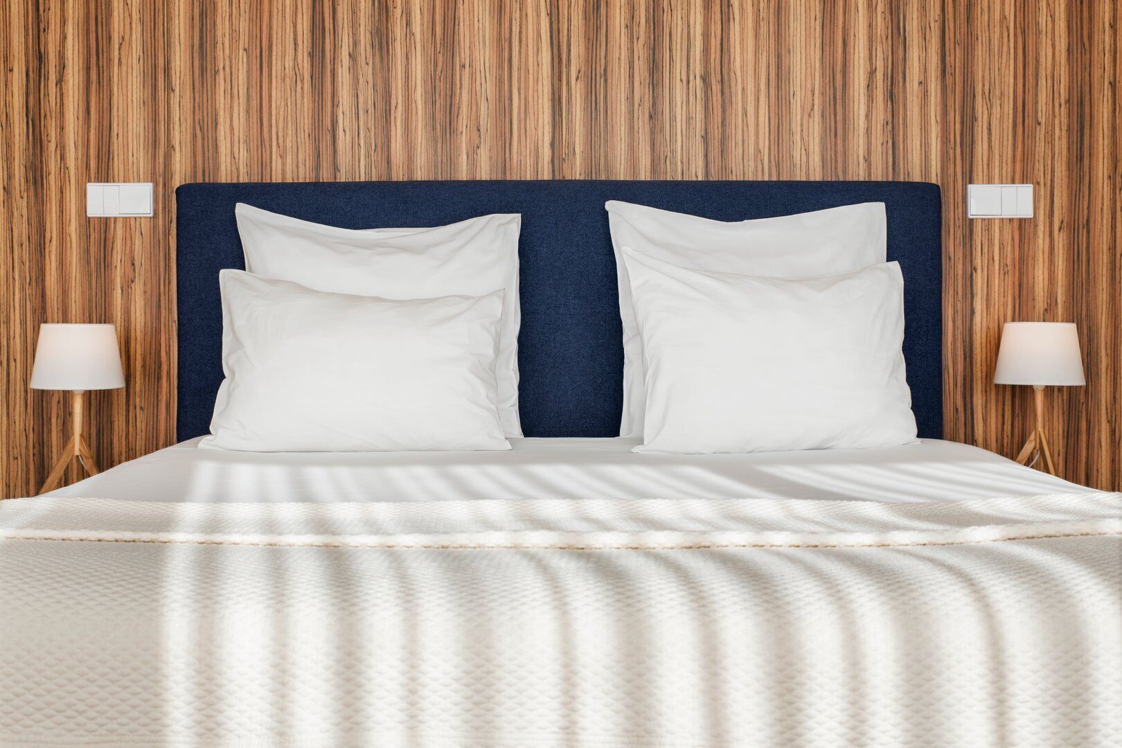 A neatly made bed with a blue headboard, white pillows, and a white duvet. There are two bedside lamps on either side of the bed.