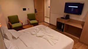 Ein Hotelzimmer mit einem ordentlich bezogenen Bett, zwei grünen Sesseln, einem an der Wand befestigten Fernseher und einem kleinen Holfnachtkästchen mit einer Kaffeemaschine.