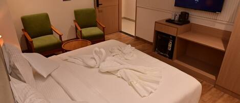 A hotel room with a neatly made bed, two green armchairs, a television on the wall, and a small wooden side table with a coffee pot.