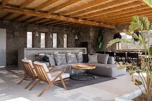 A cozy, rustic interior with a stone wall, wooden ceiling beams, a grey sofa with cushions, a wooden coffee table, and a large plant. - Villa Aspasia, pool & jacuzzi - My Rental Homes (Mykonos)