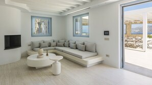 A modern living room with a L-shaped sectional sofa, a round coffee table, and a TV mounted on the wall. There are two windows providing views of the outside. - Villa Nikolas, pool and jacuzzi - My Rental Homes (Mykonos)