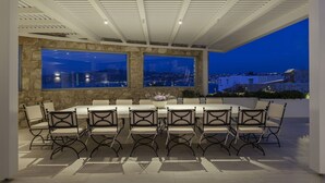An outdoor dining area with a city view at dusk, featuring a long table with chairs and a decorative centerpiece. - Villa Nikolas, pool and jacuzzi - My Rental Homes (Mykonos)
