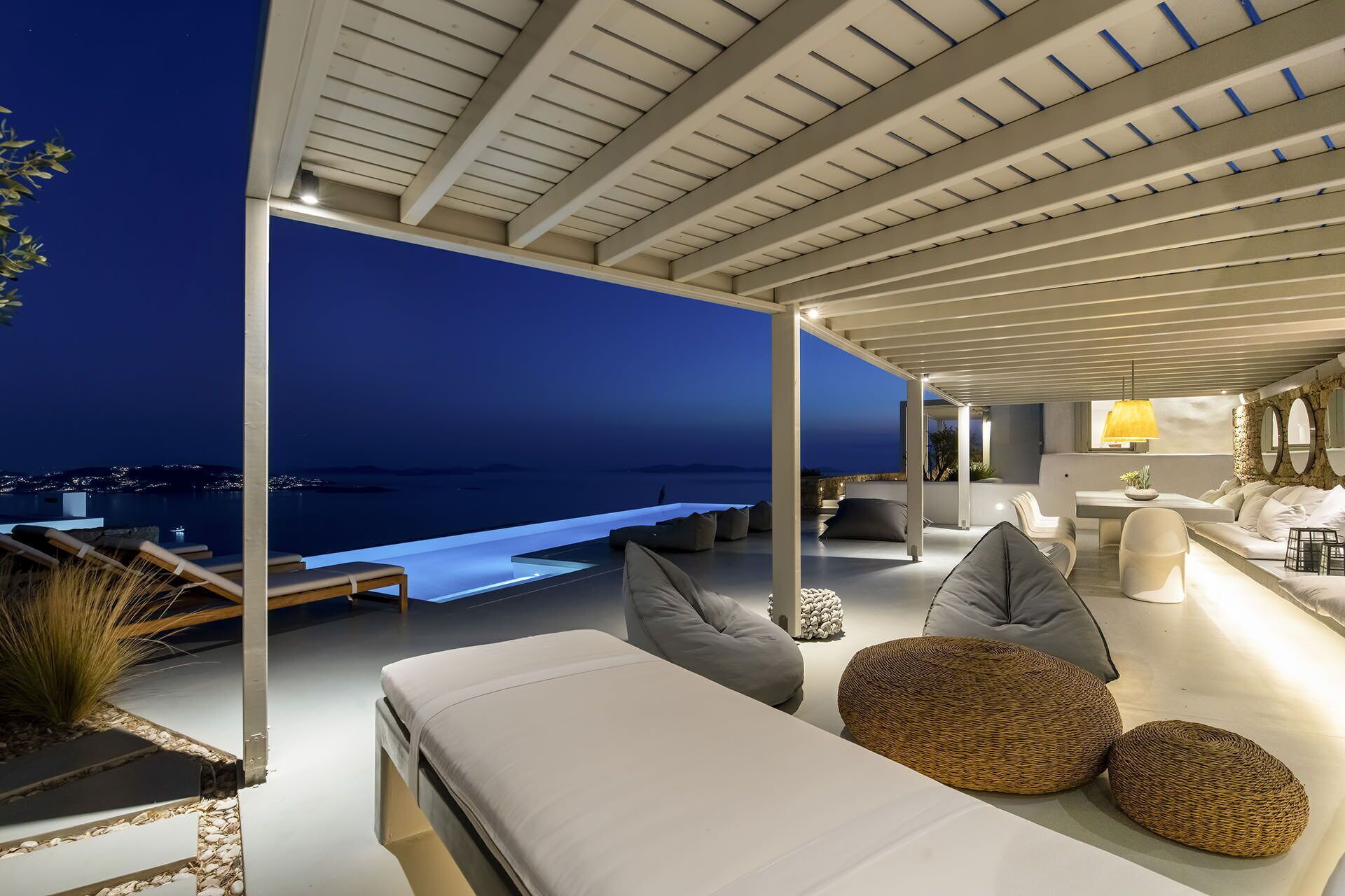 A modern outdoor lounge area with a view of the sea at night, featuring a sofa, cushions, and a dining area.