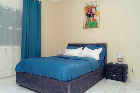 A neatly made bed with blue bedding, a nightstand with a lamp, and a wall with a colorful painting.
