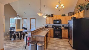 A modern kitchen with wooden cabinets, a black refrigerator, stainless steel appliances, and a dining area with a table and chairs. - 3BR Condo |  Bayfield Lake Views (Bayfield)