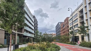 Moderni kaupunkikatu, jossa on rakennuksia, punainen pyörätie ja maisemoitu alue, jossa on kasveja ja penkki.