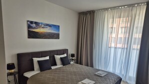 A bedroom with a large bed, a nightstand, a lamp, a painting on the wall, and a window with curtains. - Smichov Luxury apartments (Prague 5)