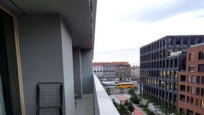 A balcony with a metal rack, overlooking a city street with buildings and a yellow bus. - Smichov Luxury apartments (Prague 5)