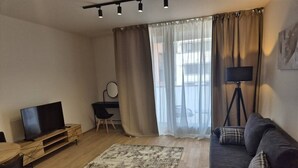 A living room with a flat-screen TV, a sofa, a coffee table, a wooden side table, a desk with a chair, and a floor lamp. - Smichov Luxury apartments (Prague 5)