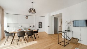 Une cuisine moderne avec des meubles blancs, un coin repas avec une table et des chaises en bois, et un téléviseur fixé au mur.