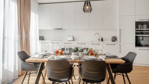 Une cuisine moderne avec des meubles blancs, une table à manger en bois et des chaises noires. On peut voir un panier de fruits et des verres à vin sur la table.