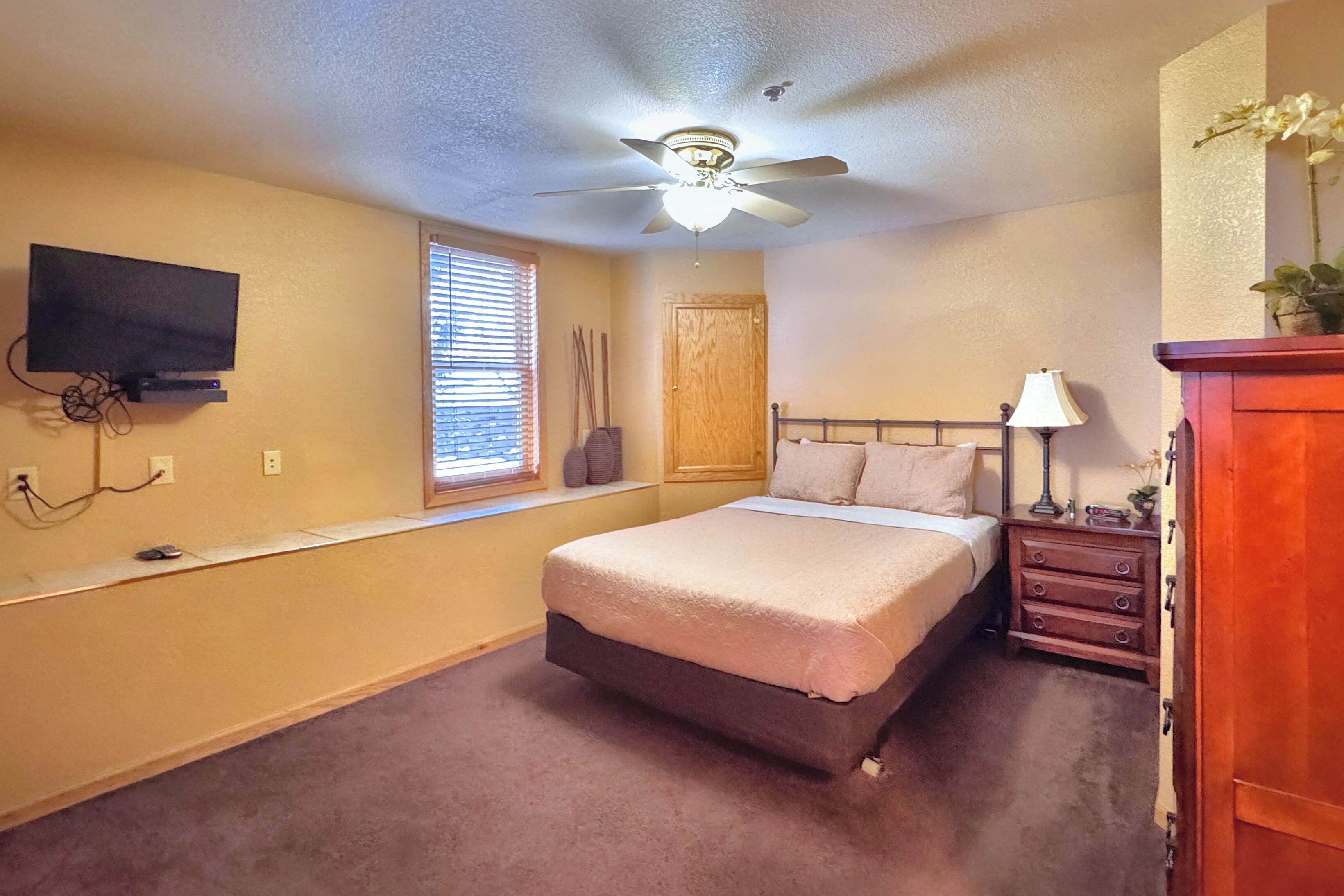 A bedroom with a bed, a ceiling fan, a TV mounted on the wall, a window with blinds, a wooden door, and a nightstand with a lamp.