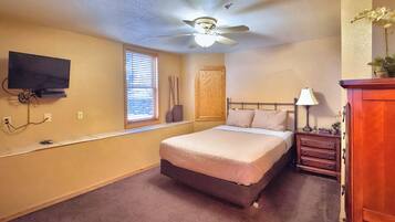 A bedroom with a bed, a ceiling fan, a TV mounted on the wall, a window with blinds, a wooden door, and a nightstand with a lamp.