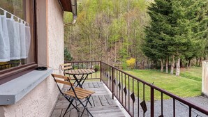 En balkon med et træbord og stole, gelænder i metal og udsigt over et skovområde.