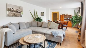 En stue med en hjørnesofa, et rundt sofabord, et spiseområde med et træbord og stole, samt en reol.