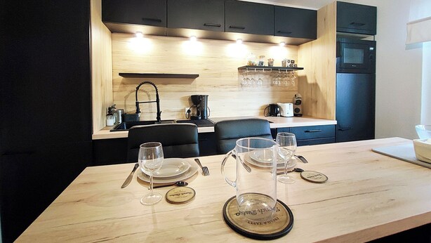 Une cuisine moderne avec des meubles de couleur noire, un plan de travail en bois et un coin repas agrémenté de verres à vin et d’assiettes.