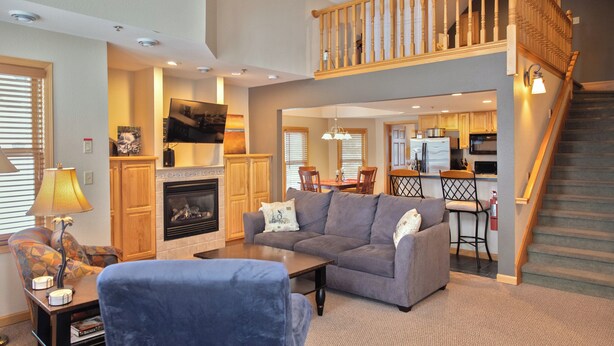 A spacious living room with a fireplace, a sofa, a coffee table, and a staircase leading to an upper level. - Top-Floor Condo | Lake Views | Walk Downtown (Bayfield)