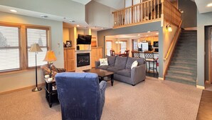 A spacious living room with a fireplace, a staircase to an upper level, and a kitchen area visible through an open door. - Top-Floor Condo | Lake Views | Walk Downtown (Bayfield)