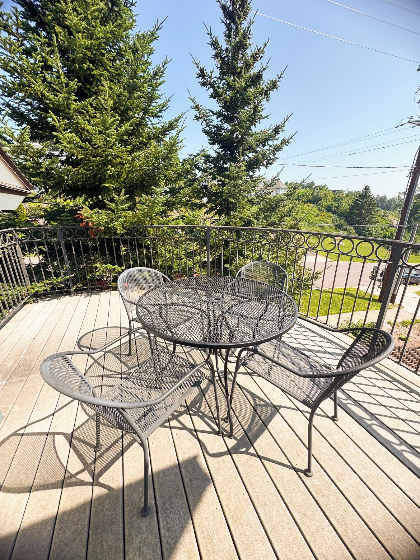 A metal outdoor table and chairs on a wooden deck with a metal fence and trees in the background.