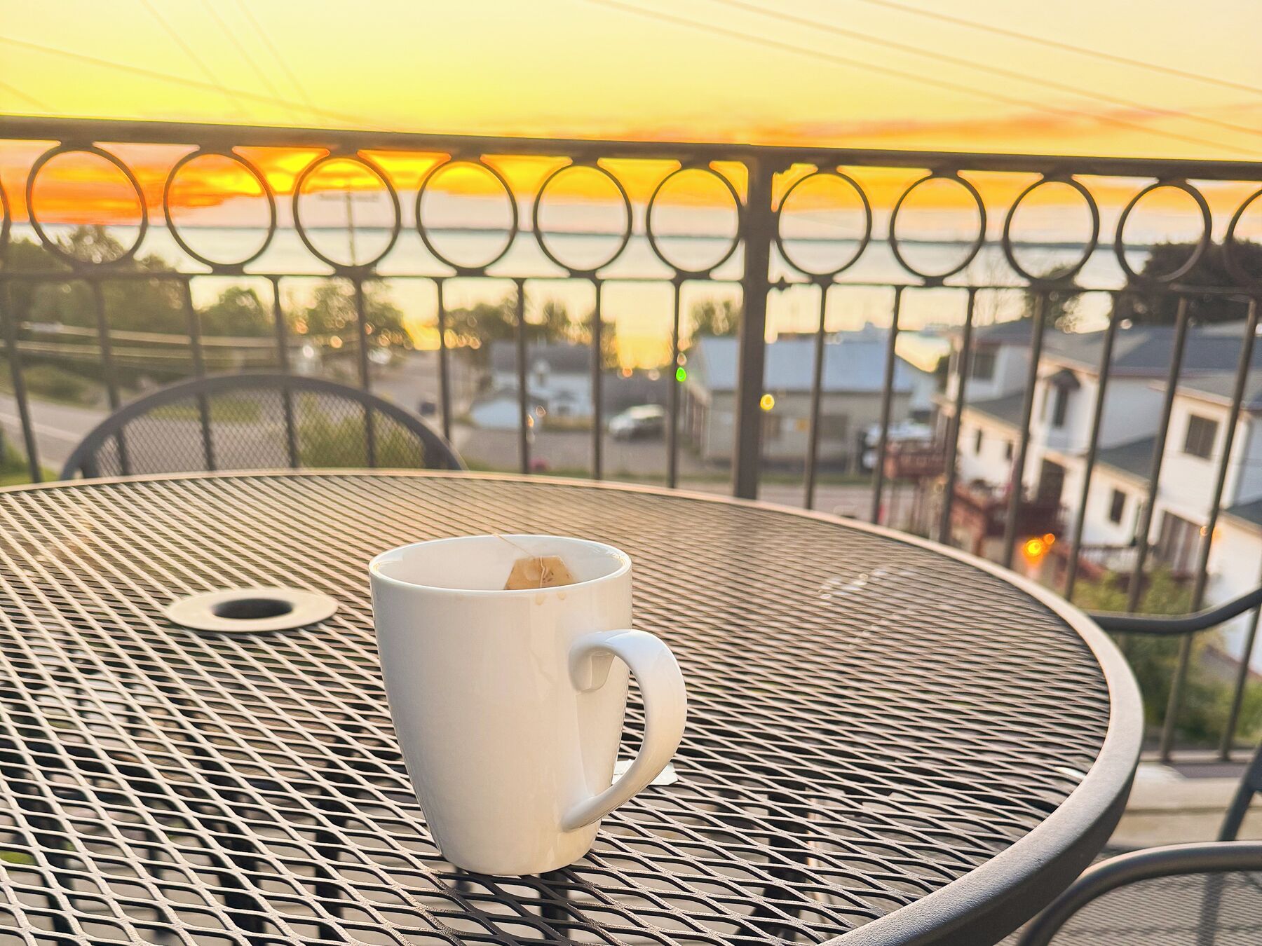 A white mug on a round metal table with a metal railing and a view of a sunset.